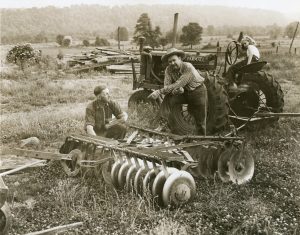 A woman sitting on a tractor and two men leaning on a piece of farm equipment and talking. They are in the middle of a field with trees in the background.