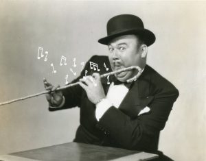 A man dressed a tuxedo, wearing a bowler hat. He is pretending to play his cane like a musical instrument. Musical notes have been added to the photo.