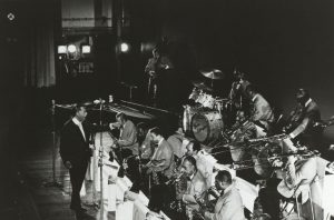Black and white image of Duke Ellington standing and conducting his 11 piece ensemble known as the Washingtonians.