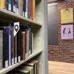 photograph of books shelved in Sawyer library with a bookmark with a black geolocation pin on it.