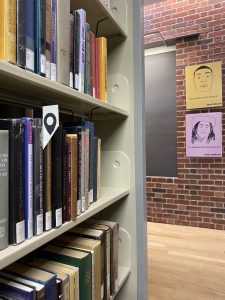 photograph of books shelved in Sawyer library with a bookmark with a black geolocation pin on it.