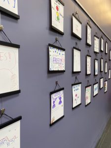 side view of a purple wall with white drawings framed in black hanging in a display.